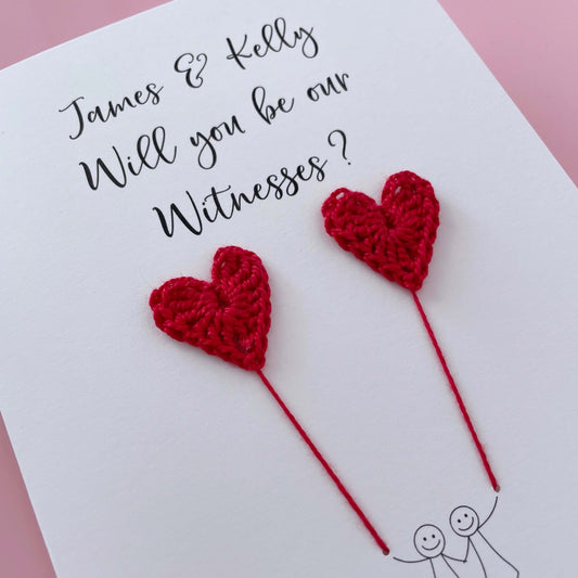 A white card with a line drawn couple printed on it - the couple are each holding a Red crochet heart balloon. Will you be our witnesses is printed at the top of the card with the option of adding a name, making it the perfect keepsake. 
