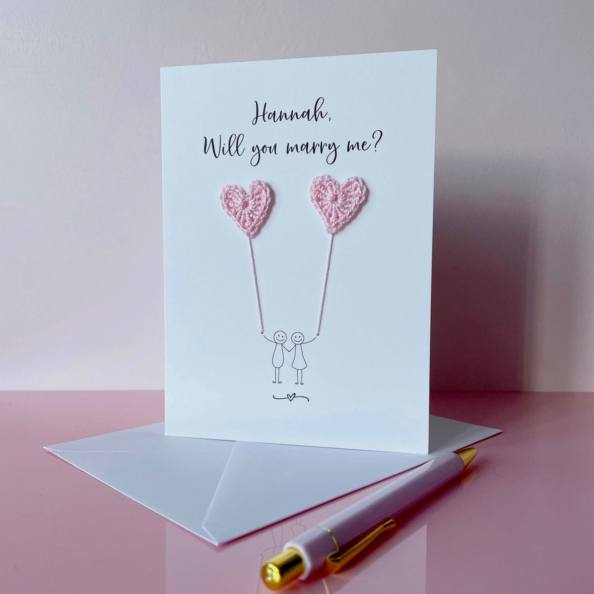 Greeting card with crochet heart-shaped balloons and text 'Hannah, Will you marry me?' on a pink surface with a pen.