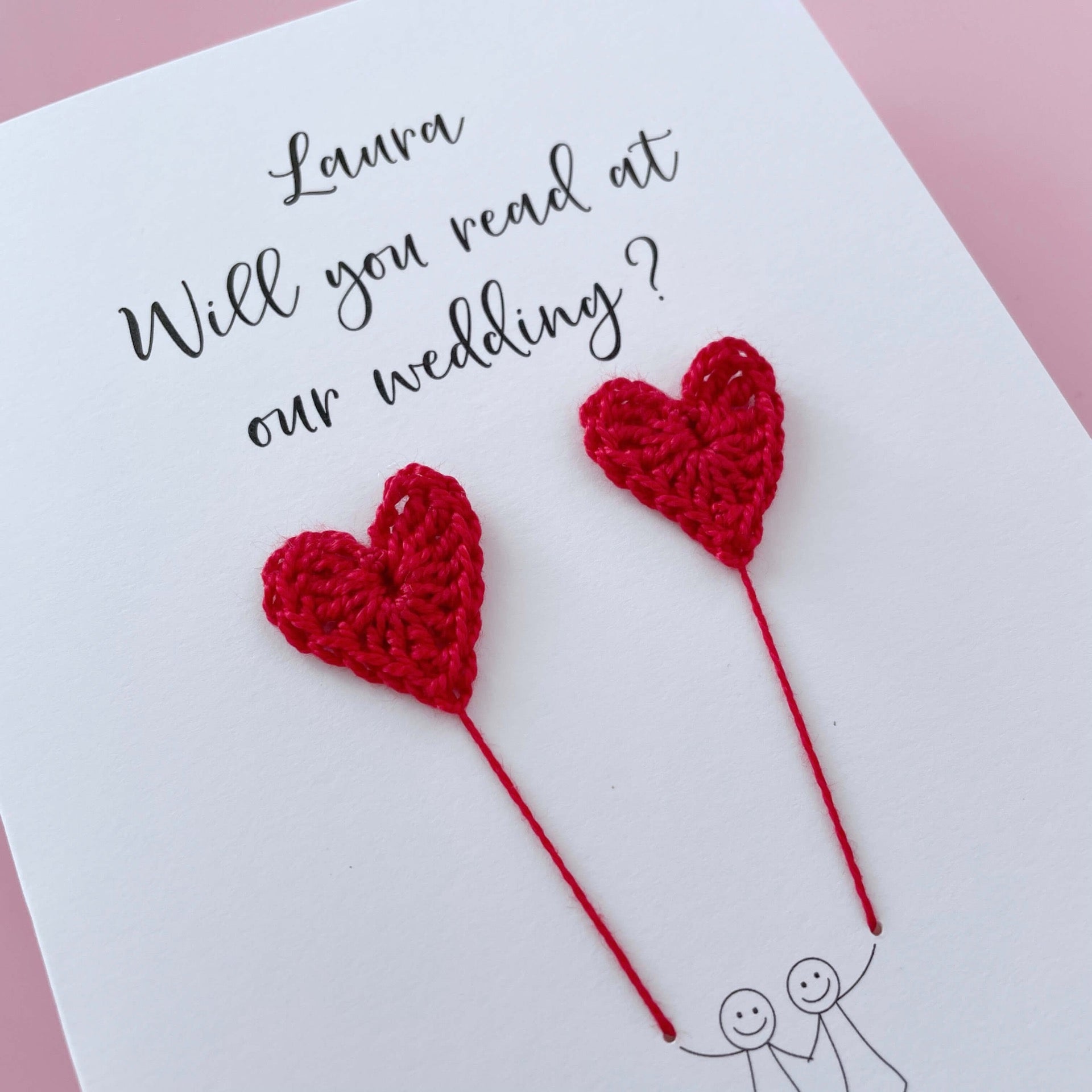 A white card with a line drawn couple printed on it - the couple are each holding a red crochet heart balloon. Will you read at our wedding is printed at the top of the card with the option of adding a name, making it the perfect keepsake. 