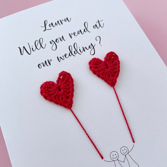 A white card with a line drawn couple printed on it - the couple are each holding a red crochet heart balloon. Will you read at our wedding is printed at the top of the card with the option of adding a name, making it the perfect keepsake. 