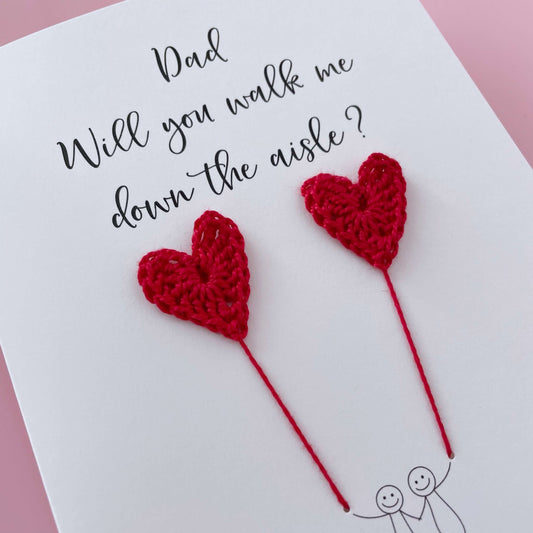 A white card with a line drawn couple printed on it - the couple are each holding a Red crochet heart balloon. Will you walk me down the aisle is printed at the top of the card with the option of adding a name, making it the perfect keepsake. 