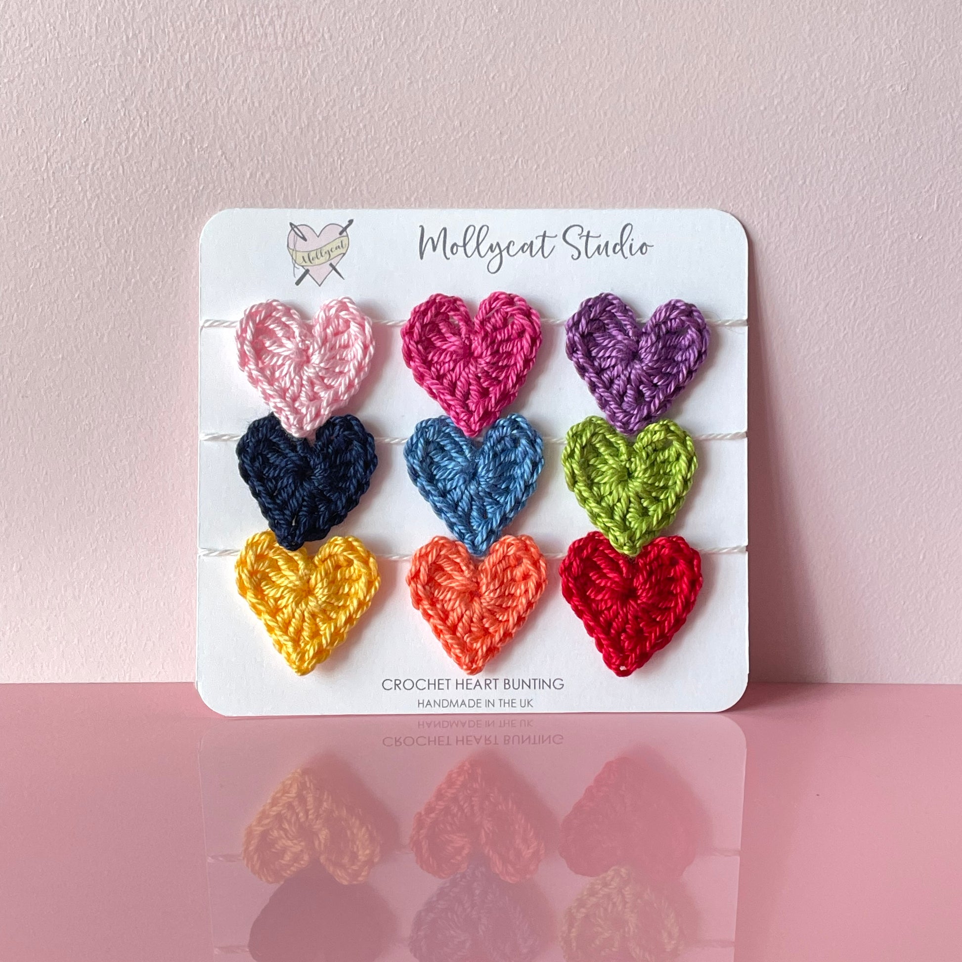 Mini crochet heart bunting. 9 mini crochet hearts in the colours of the rainbow, strung onto white cotton yarn. The bunting is packaged on white card, placed on a pale pink background.