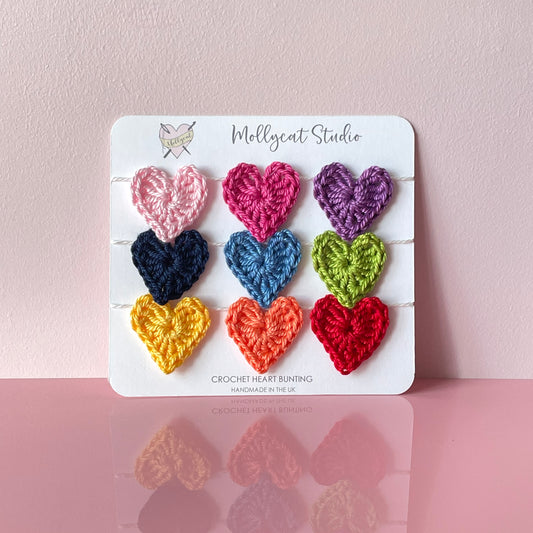 Mini crochet heart bunting. 9 mini crochet hearts in the colours of the rainbow, strung onto white cotton yarn. The bunting is packaged on white card, placed on a pale pink background.