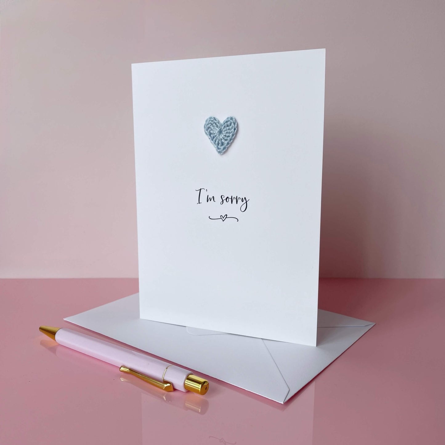A white card with I'm sorry printed in the centre of it in black cursive font. A pale blue crochet heart is on the card above the text. The card is on a palace pink background.