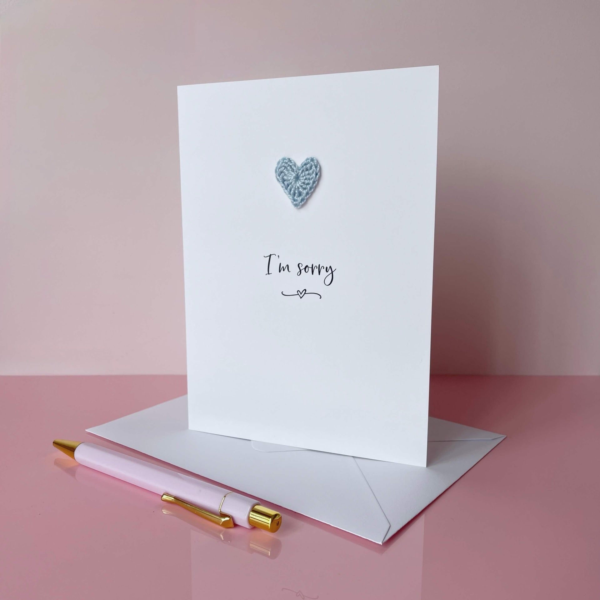 A white card with I'm sorry printed in the centre of it in black cursive font. A pale blue crochet heart is on the card above the text. The card is on a palace pink background.