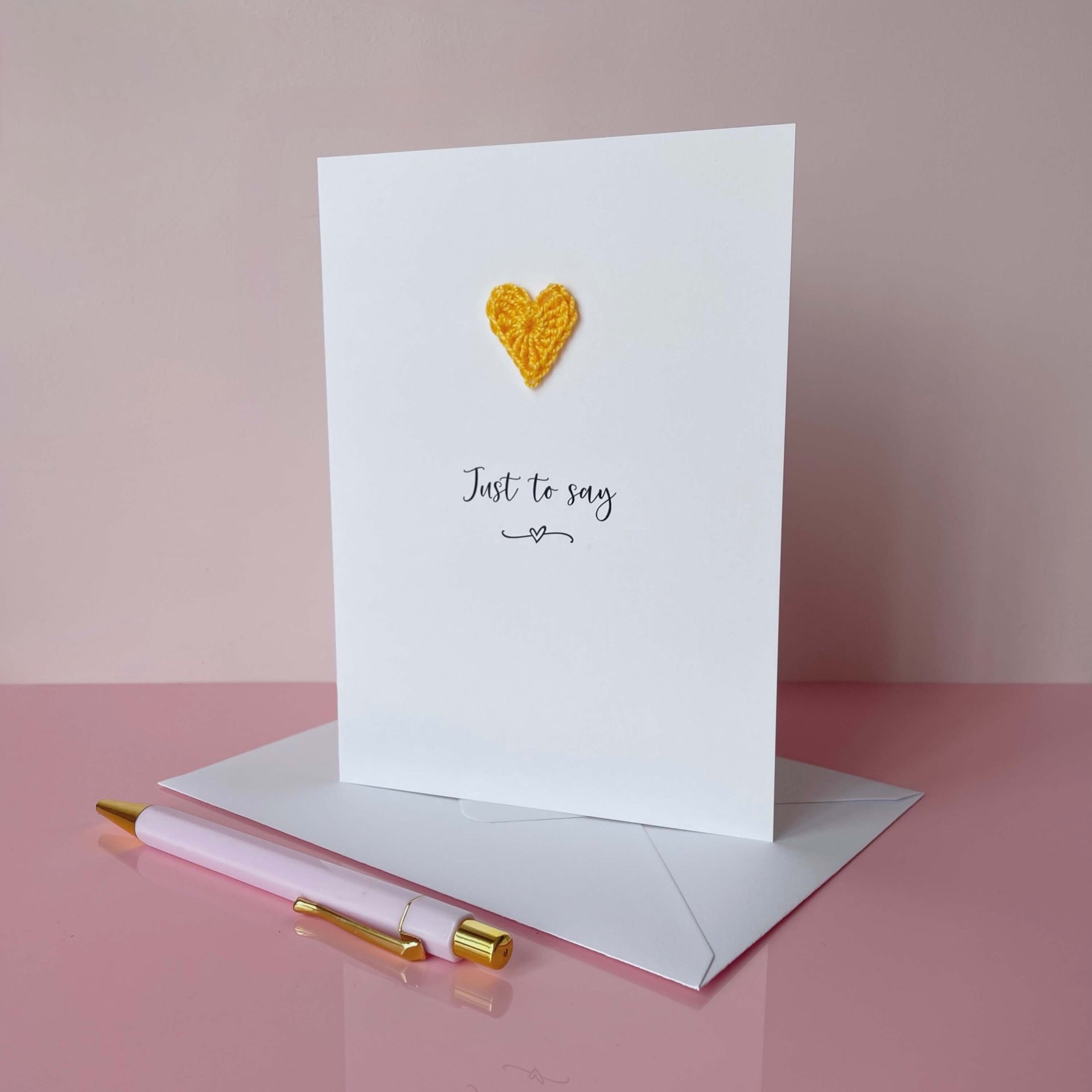 A white card with Just to say printed in the centre of it in black cursive font. A yellow crochet heart is on the card above the text. The card is on a placed pink background.