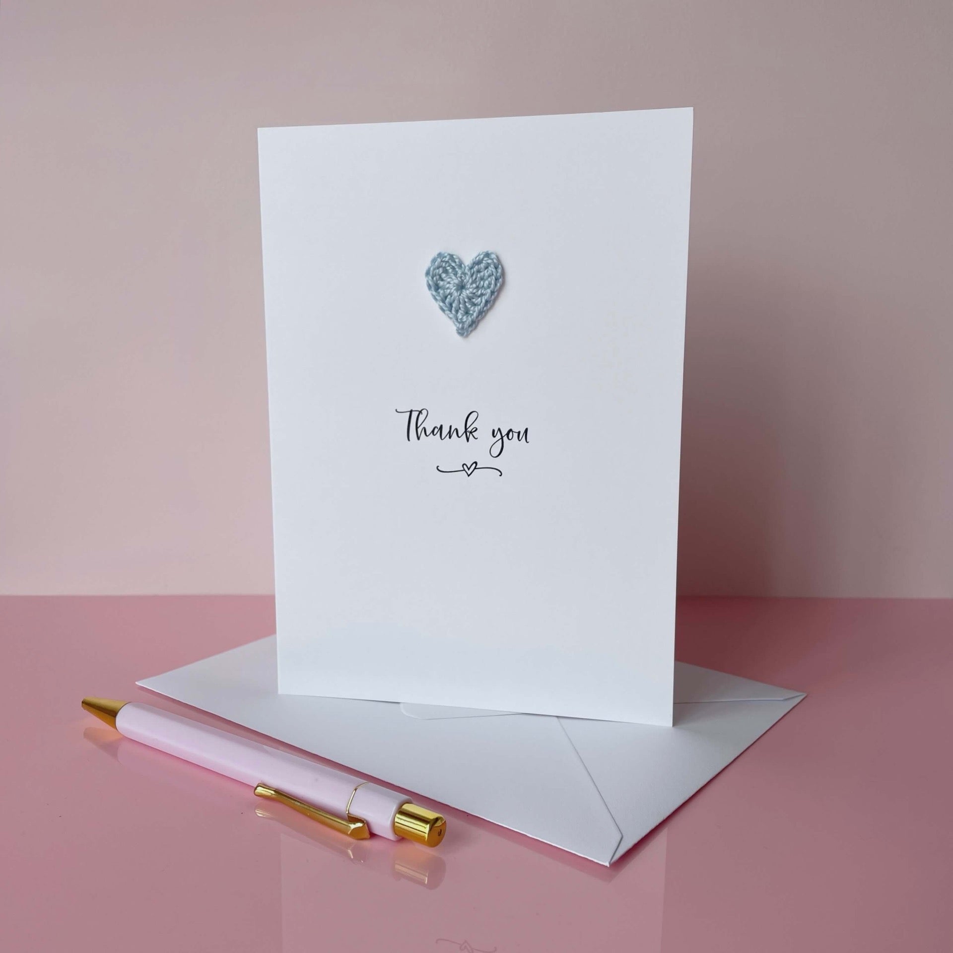 White card with a heart design and 'Thank you' text, accompanied by a pink envelope and pen on a pink background.