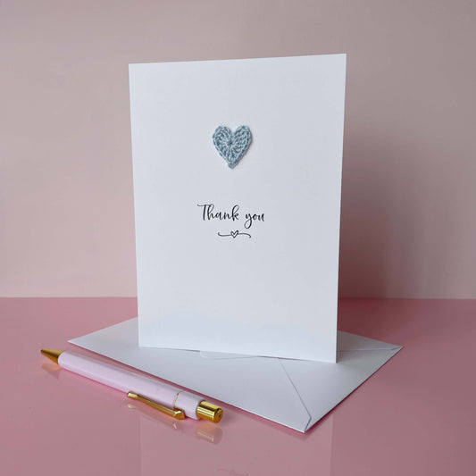 White card with a heart design and 'Thank you' text, accompanied by a pink envelope and pen on a pink background.