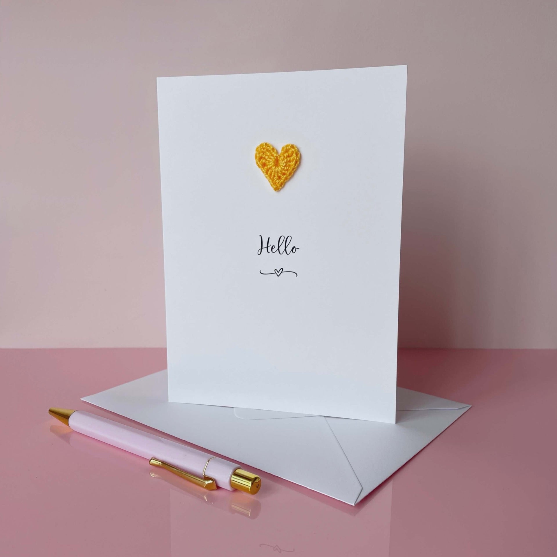 Greeting card with yellow heart and 'Hello' text on a pink surface with a pen and envelope.