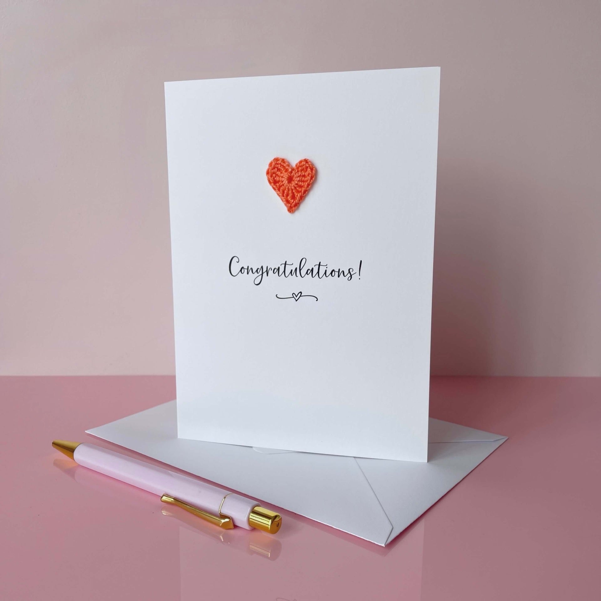 A white card with Congratulations! printed in the centre of it in black cursive font. A coral coloured crochet heart is on the card above the text. The card is on a placed pink background.