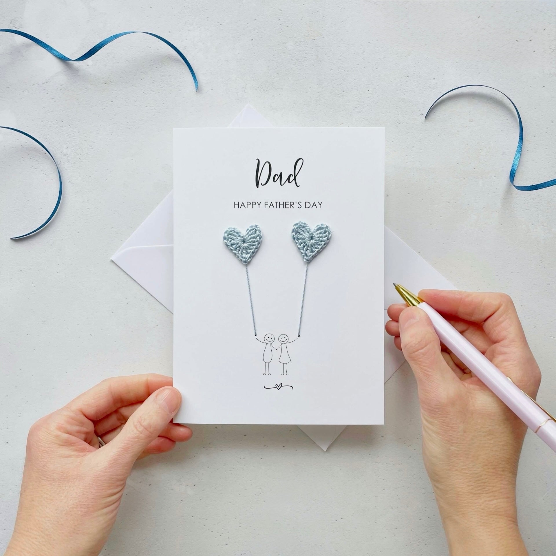 A simple father's day card with crochet hearts featuring a minimalist design with two line drawn figures holding pale blue crochet heart balloons. The text at the top reads: ‘Dad, Happy Father’s Day. 
