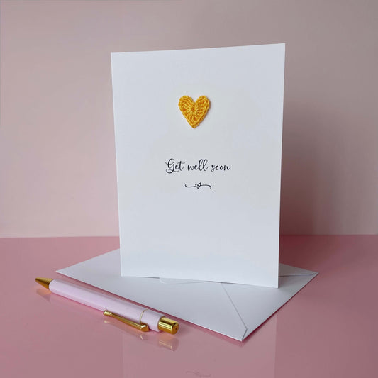 A white card with get well soon printed in the centre of it in black cursive font. A yellow crochet heart is on the card above the text. The card is on a placed pink background.