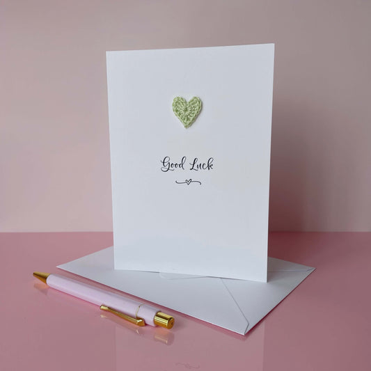 A white card with Good luck printed in the centre of it in black cursive font. A pale green crochet heart is on the card above the text. The card is on a placed pink background.