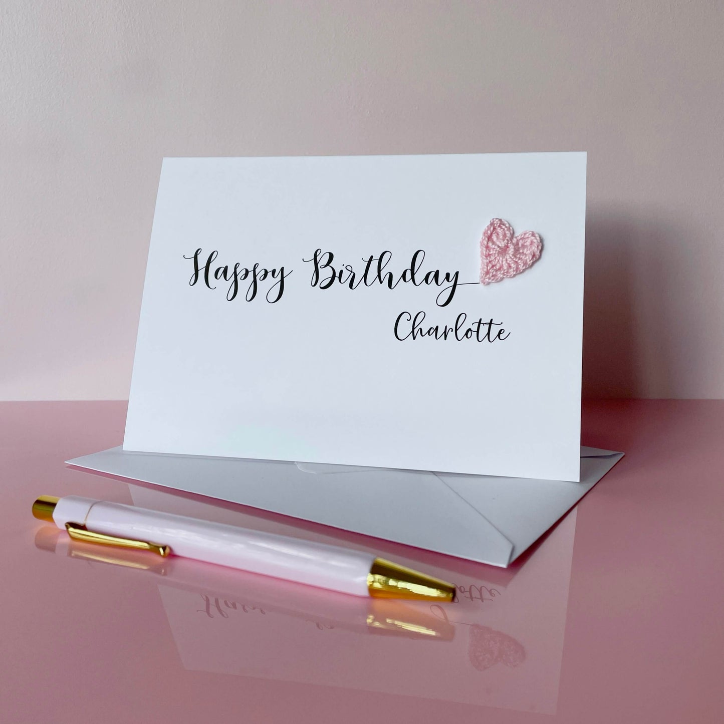 A white birthday card with 'Happy Birthday Charlotte' printed in black cursice text. A pale pink crochet heart has been attatched to the 'y' at the end of Birthday. 