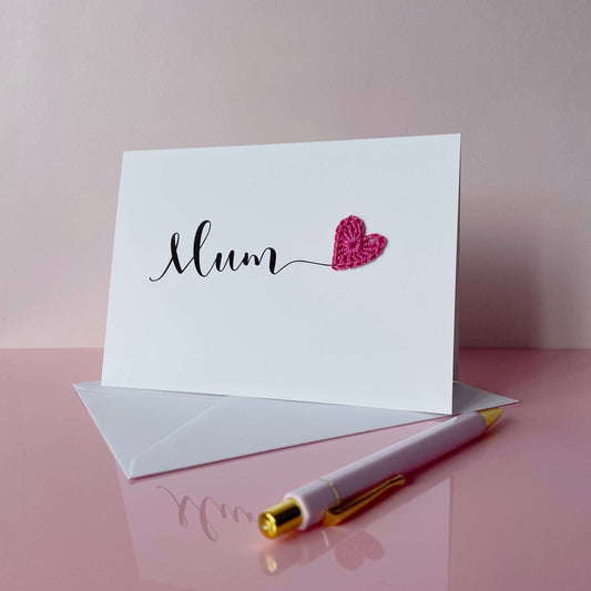 A white card with the word 'Mum' printed across the middle in black script text. There is a bright pink crochet heart stuck onto the end of the word 'Mum'
