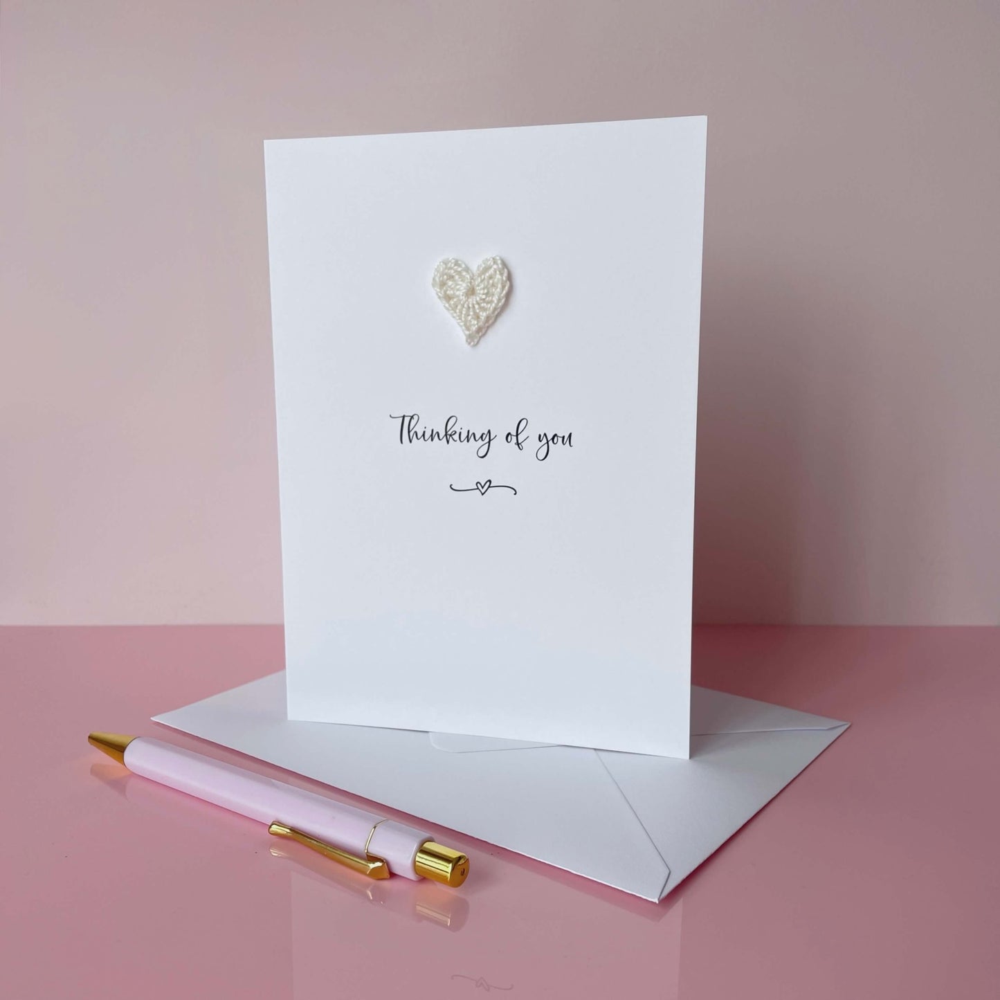 A white card with Thinking of you printed in the centre of it in black cursive font. A white crochet heart is on the card above the text. The card is on a placed pink background.