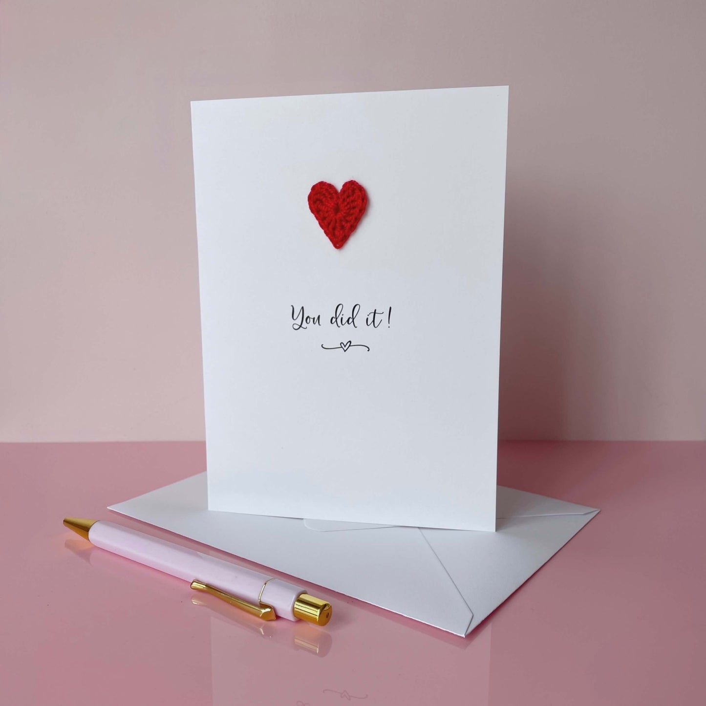 Greeting card with red heart and 'You did it!' text on a pink surface with a pen and envelope.