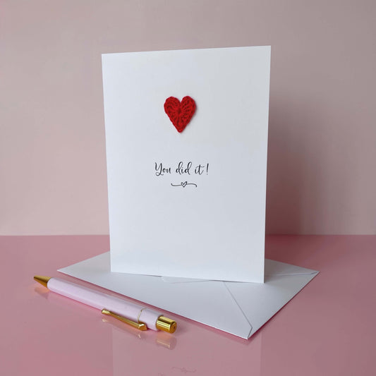 Greeting card with red heart and 'You did it!' text on a pink surface with a pen and envelope.