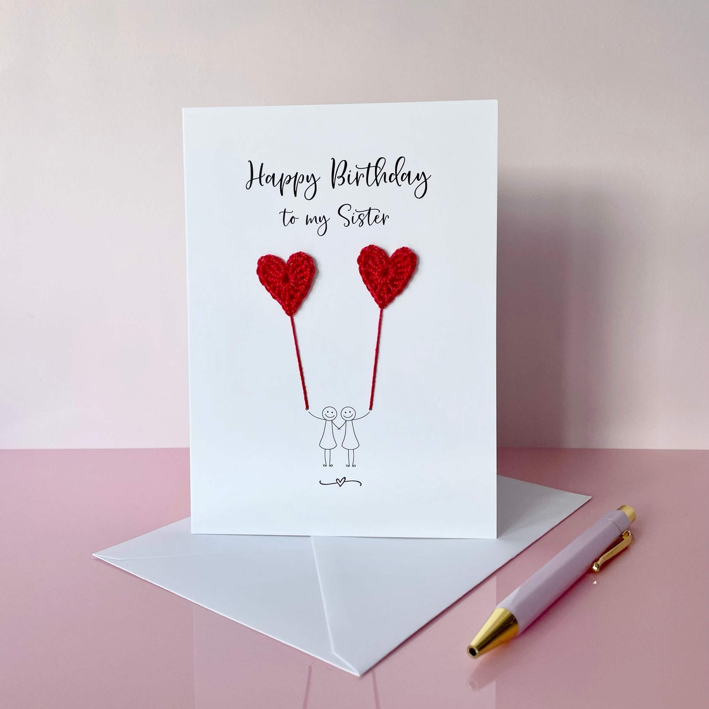 happy birthday to my Sister is printed at the top of the card and a line drawn couple are each holding a red crochet heart balloon. 
