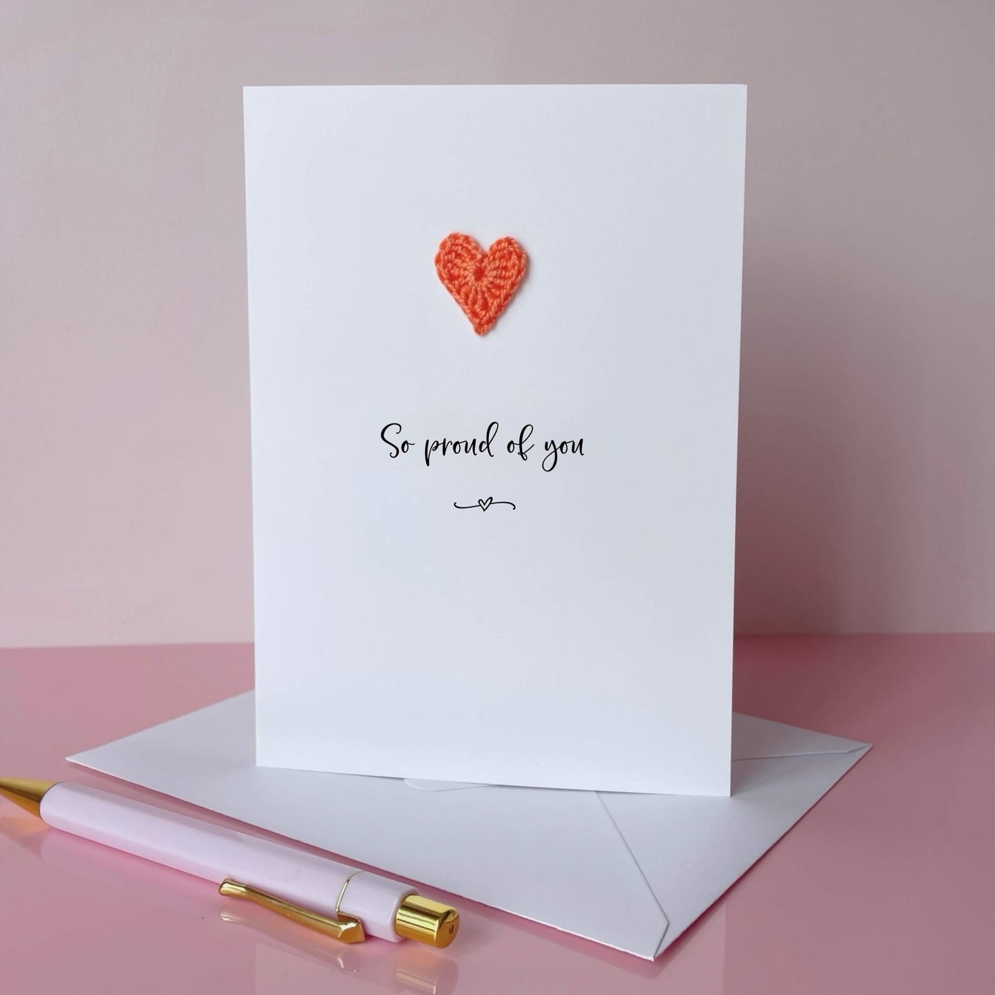 Greeting card with a crochet heart and text on a pink background, 'So proud of you' is printed on the card in lovely black cursive text. 