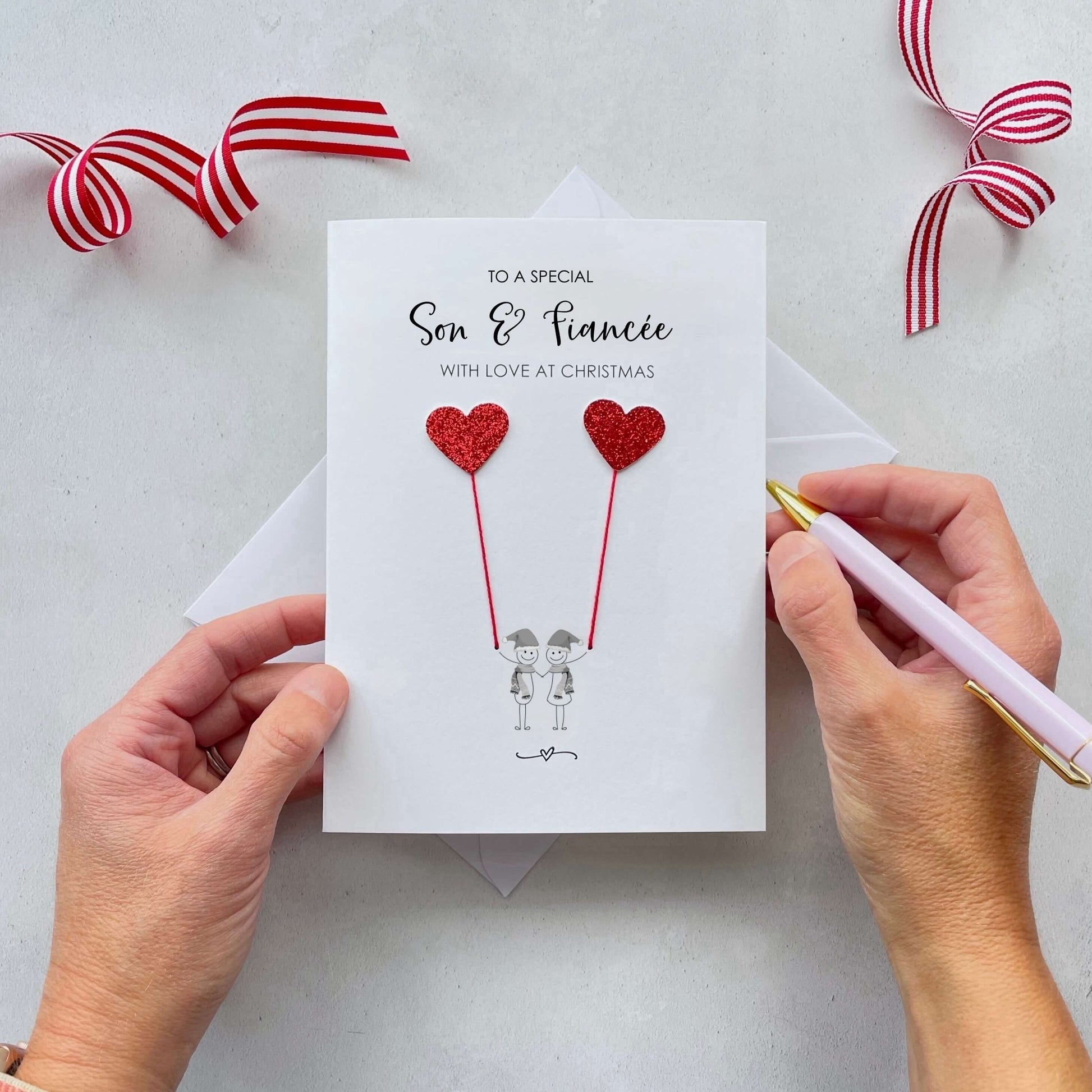 A Christmas card featuring a minimalist design with two figures wearing santa hats holding red glitter heart balloons. The text at the top reads: ‘To a special Son & fiancée, with love at Christmas’. The card is placed on a white background with a white envelope partially visible behind it. A hand is seen holding the card, while another hand holds a pink pen. A red and white striped ribbon is positioned in the top corners of the image for decoration.