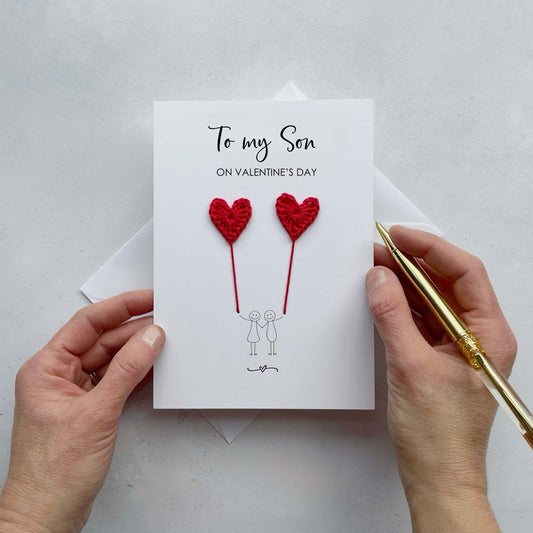 A white card featuring a line drawn couple holding hands and each holding a red crochet heart balloon.  ‘To my Son on Valentine’s Day’ is printed at the top of the card in black cursive text.