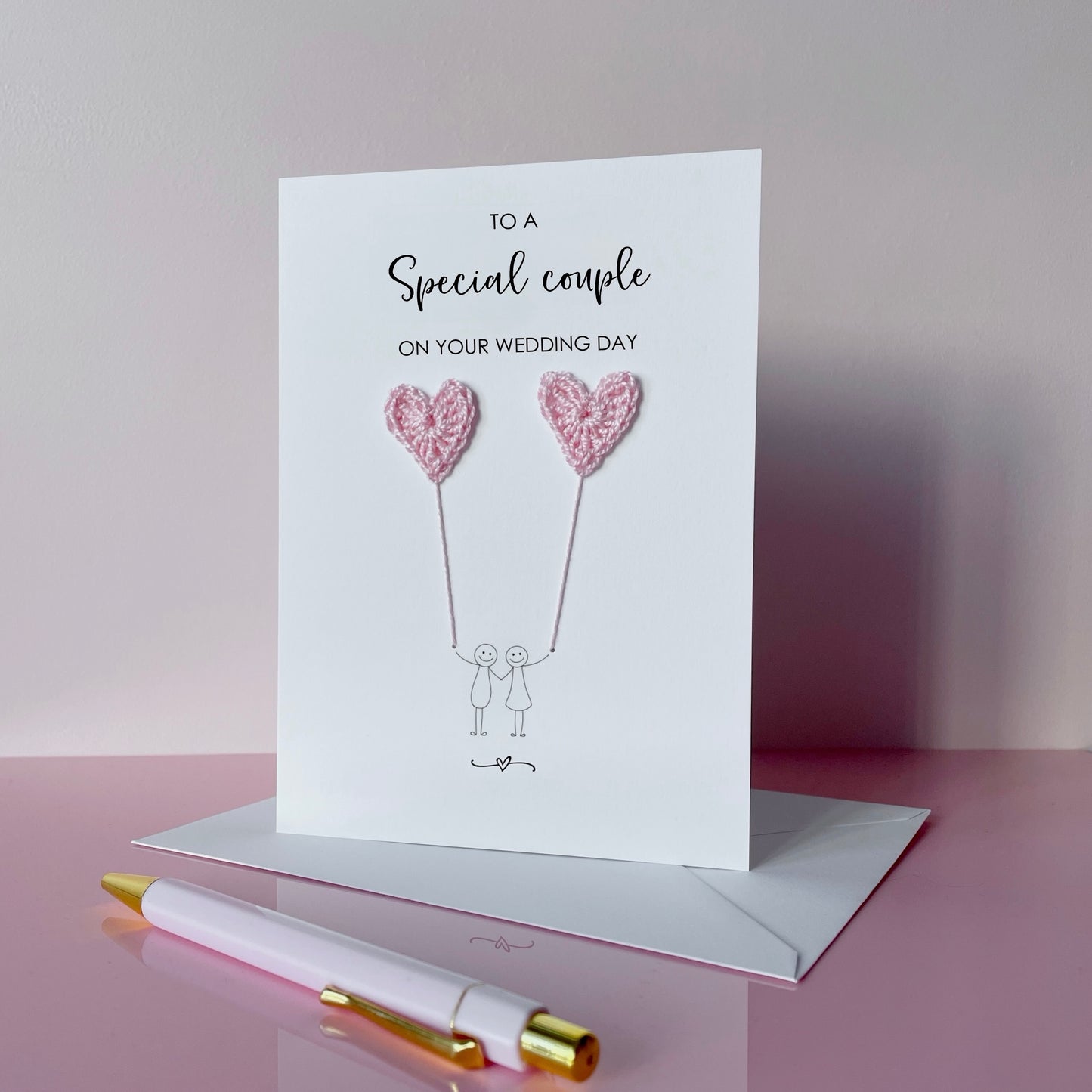A white greeting card with pale pink crochet heart balloons and text on a pink surface. The text at the top of the cad reads 'To a special couple on your wedding day' 