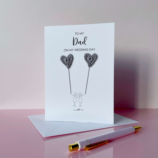 A white (portrait) card with ‘To my Dad on my wedding day’ printed at the top of in black cursive text. 
A line drawn couple holding hands is printed on the card, they are each holding a grey crochet heart balloon. 