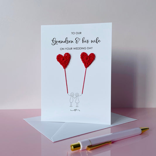 A white greeting card with red crochet heart balloons and text on a pink surface. The text at the top of the cad reads 'To our Grandson & his wife on your wedding day' 