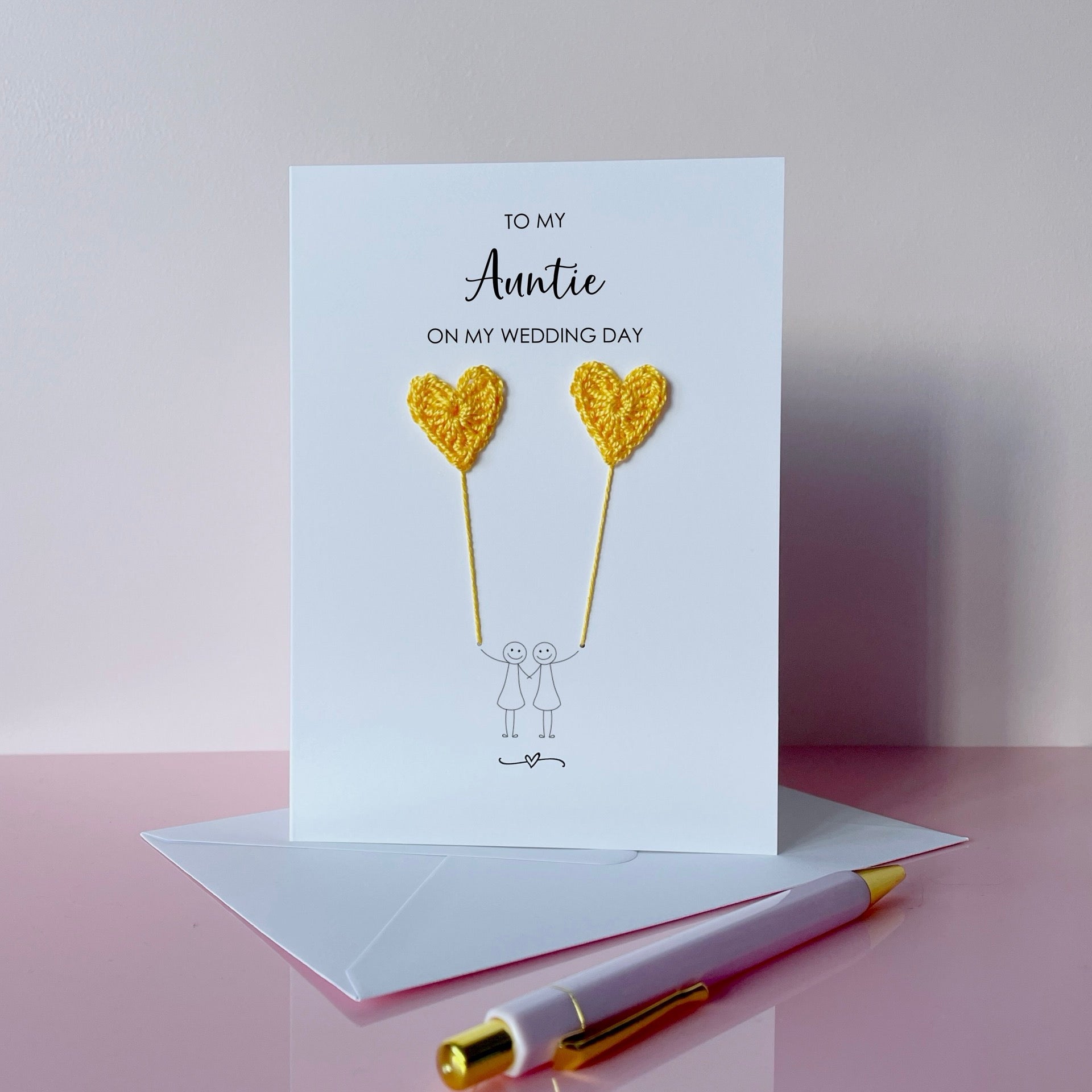 Greeting card with yellow crochet heart balloons and text on a pink surface. The text at the top of the card reads 'To my Auntie on my wedding day' 