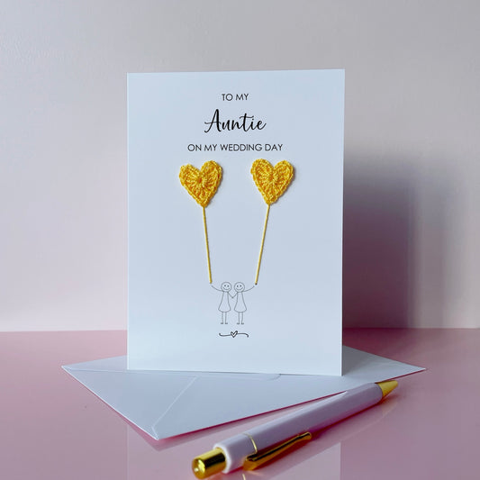 Greeting card with yellow crochet heart balloons and text on a pink surface. The text at the top of the card reads 'To my Auntie on my wedding day' 