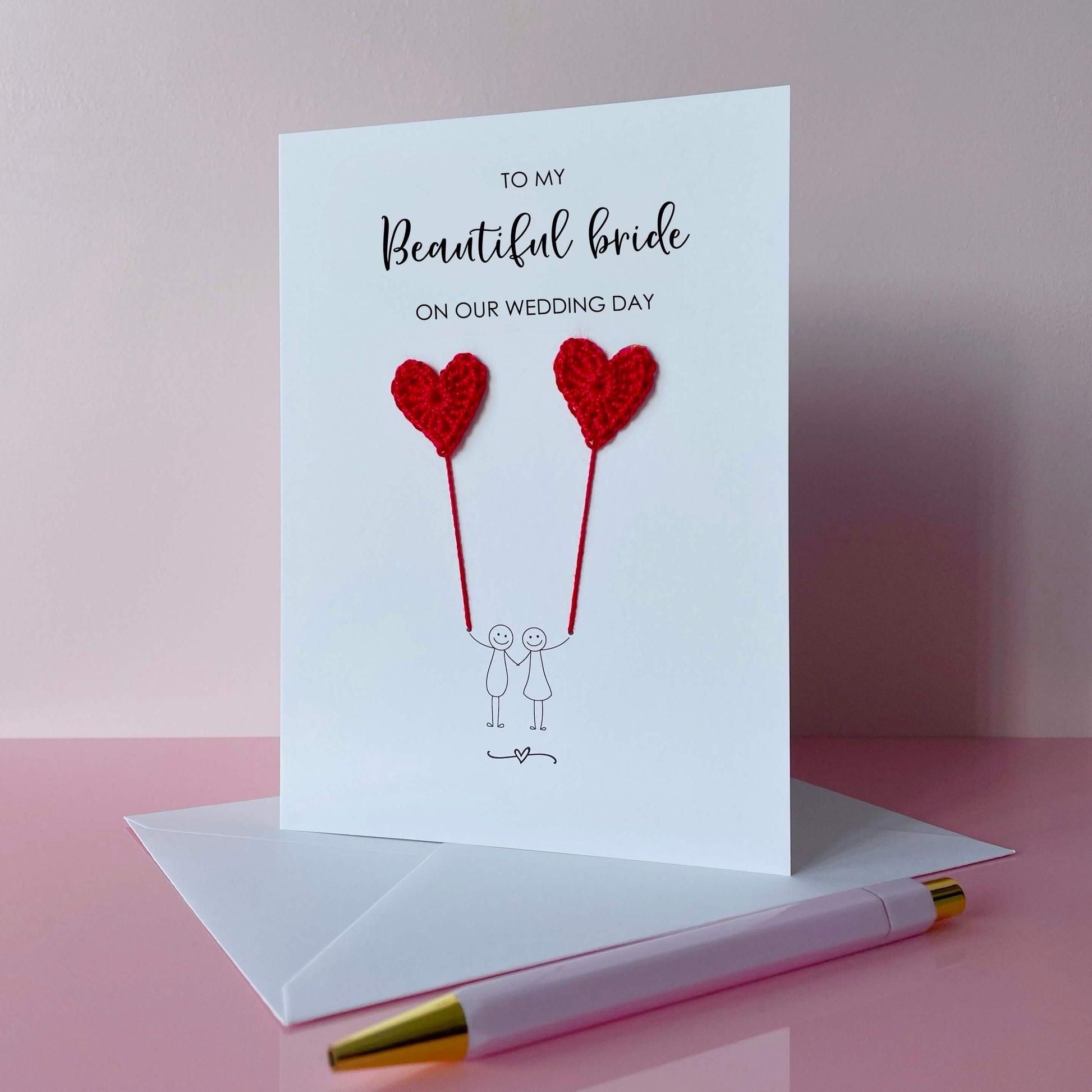 A white greeting card with a line drawn couple each holding a red crochet heart balloon. The text at the top of the card reads 'To my beautiful bride on our wedding day' 