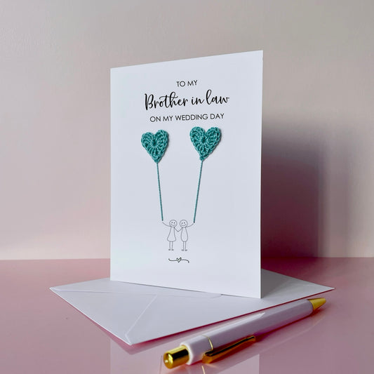 Greeting card with pale blue crochet heart balloons and text on a pink surface. The text at the top of the card reads 'To my Brother in law on my wedding day' 