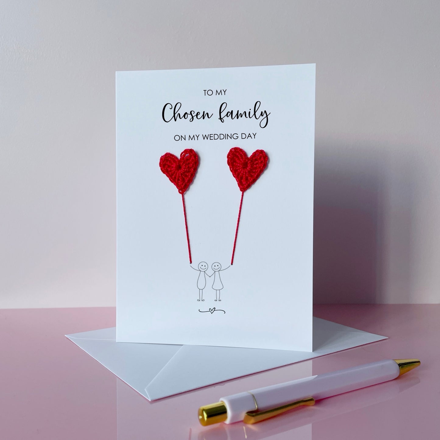 A white greeting card with red crochet heart balloons and text on a pink surface. The text at the top of the cad reads 'To my chosen family on my wedding day' 
