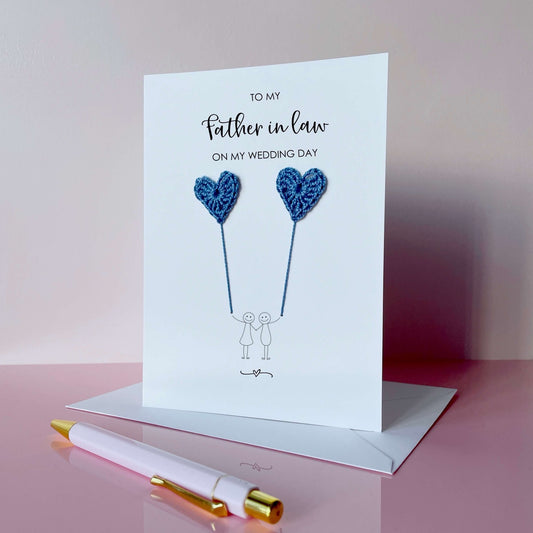 A white (portrait) card with ‘To my Father-in-law on my wedding day’ printed at the top of in black cursive text. 
A line drawn couple holding hands is printed on the card, they are each holding a blue crochet heart balloon. 