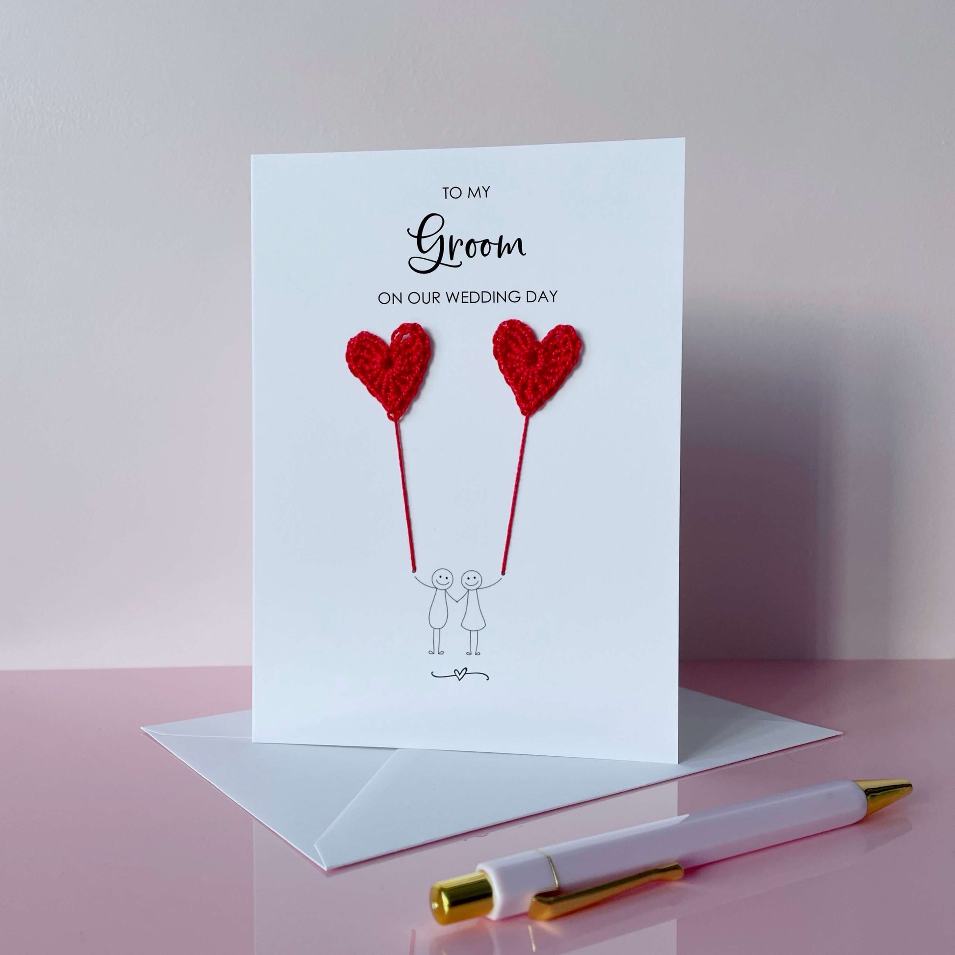 Greeting card with red crochet heart balloons and text on a pink surface. The text at the top of the cad reads 'To my Groom on our wedding day' 