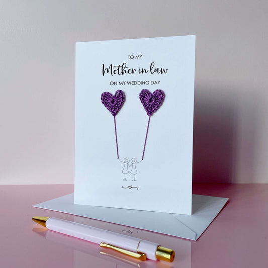 A white (portrait) card with ‘To my Mother-in-law on my wedding day’ printed at the top of in black cursive text. 
A line drawn couple holding hands is printed on the card, they are each holding a purple crochet heart balloon. 