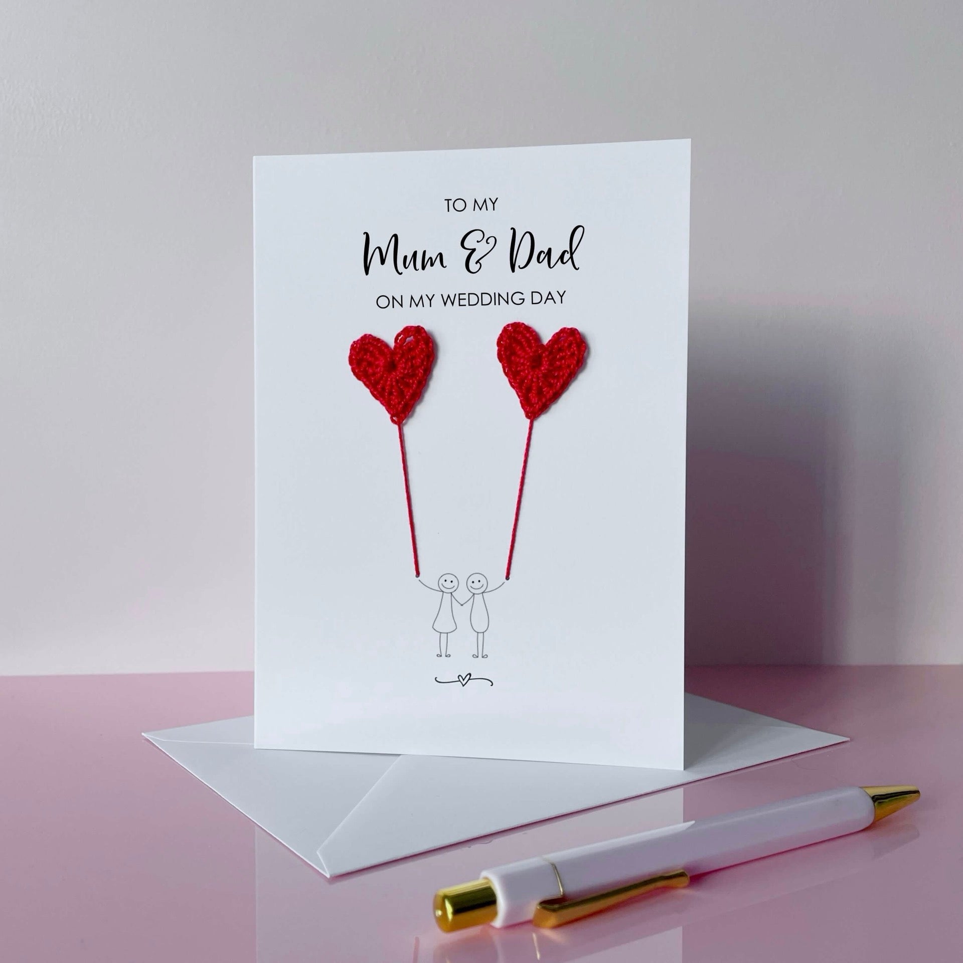 A white greeting card with red crochet heart balloons and text on a pink surface. The text at the top of the cad reads 'To my Mum & Dad on my wedding day' 