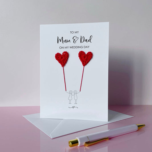A white greeting card with red crochet heart balloons and text on a pink surface. The text at the top of the cad reads 'To my Mum & Dad on my wedding day' 