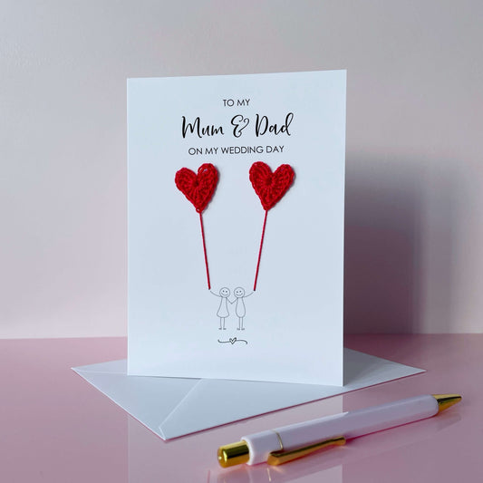 Greeting card with pink crochet heart balloons and text on a pink surface. The text at the top of the cad reads 'To my Mum & Dad on my wedding day' 