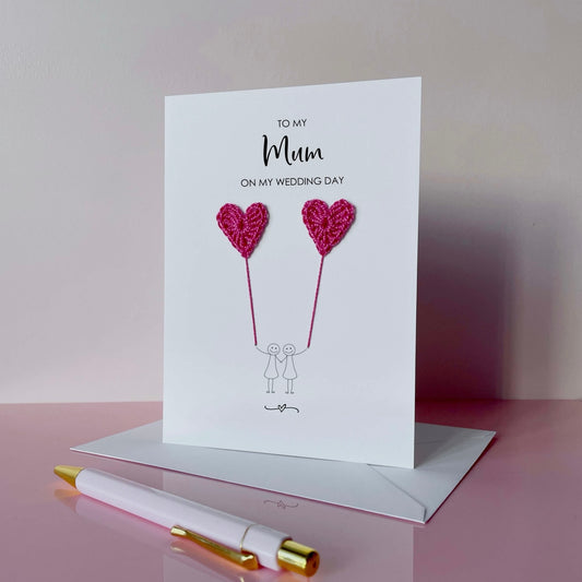 Greeting card with bright pink crochet heart balloons and text on a pink surface. The text at the top of the cad reads 'To my Mum on my wedding day' 
