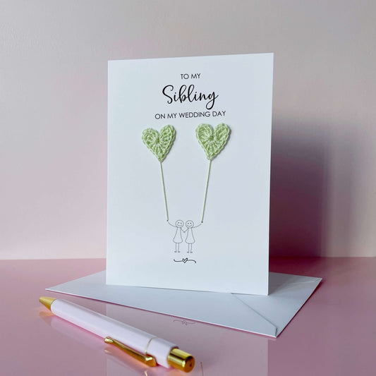 Greeting card with pale green crochet heart balloons and text on a pink surface. The text at the top of the cad reads 'To my Sibling on my wedding day' 