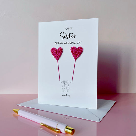 Greeting card with bright pink crochet heart balloons and text on a pink surface. The text at the top of the card reads 'To my Sister on my wedding day' 