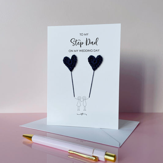 A white (portrait) card with ‘To my Step Dad on my wedding day’ printed at the top of in black cursive text. 
A line drawn couple holding hands is printed on the card, they are each holding a navy crochet heart balloon. 