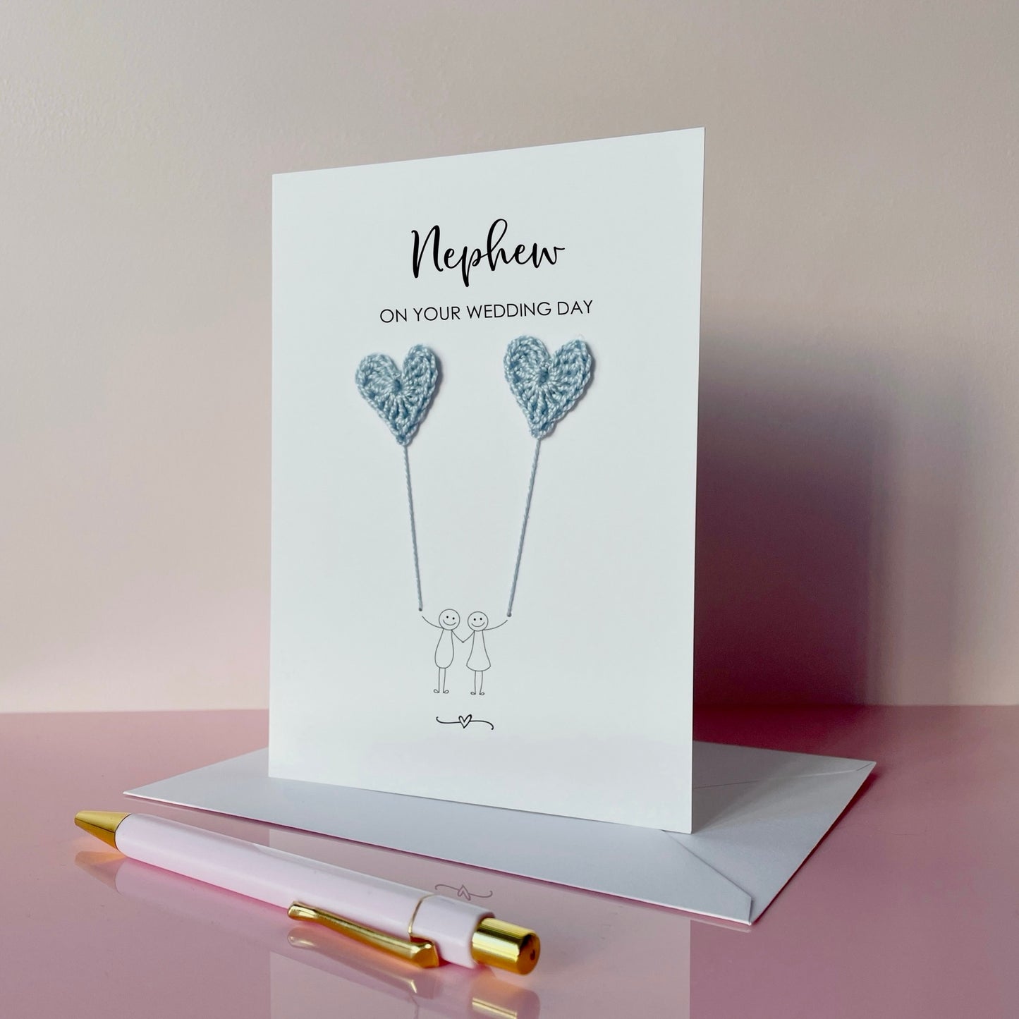 Greeting card for nephew on wedding day with pale blue heart balloons and couple illustration, placed on a pink surface with a pen.