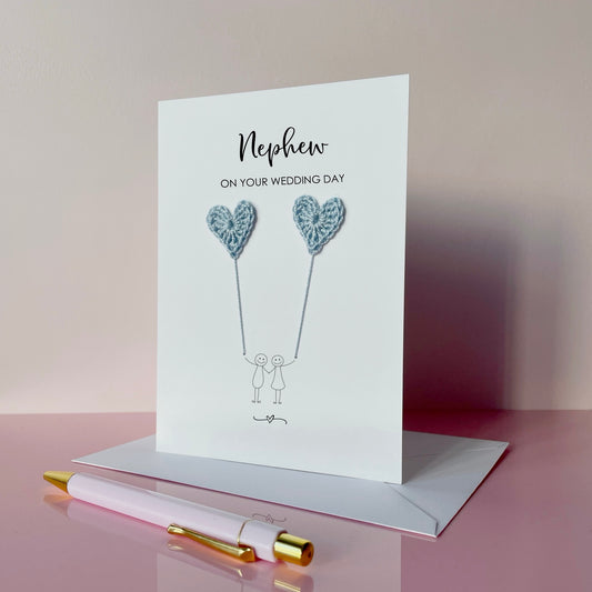 Greeting card for nephew on wedding day with pale blue heart balloons and couple illustration, placed on a pink surface with a pen.