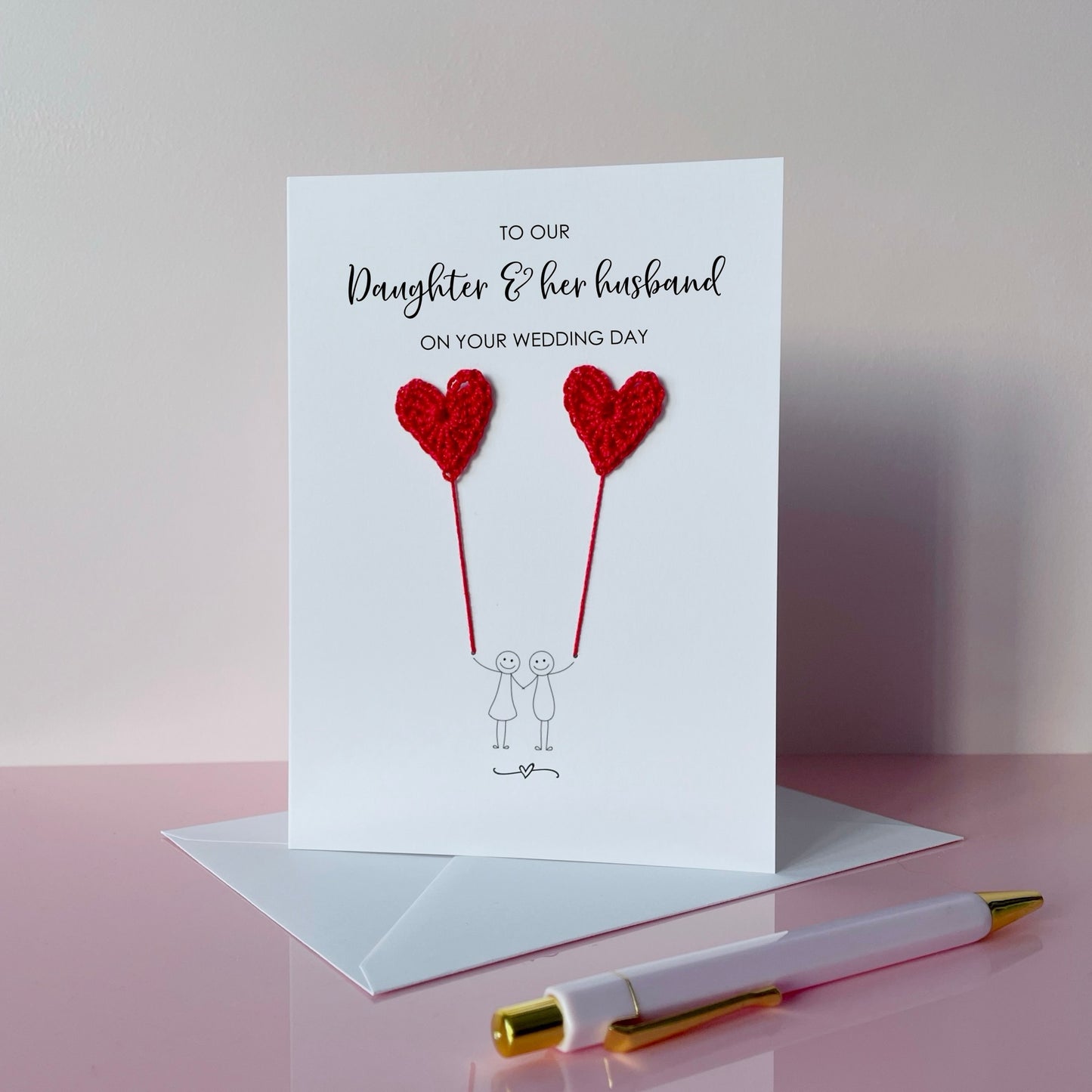 Greeting card with red crochet heart balloons and text on a pink surface. The text at the top of the cad reads 'To our Daughter & her husband on your wedding day' 