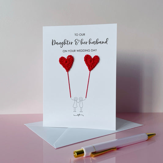 Greeting card with red crochet heart balloons and text on a pink surface. The text at the top of the cad reads 'To our Daughter & her husband on your wedding day' 
