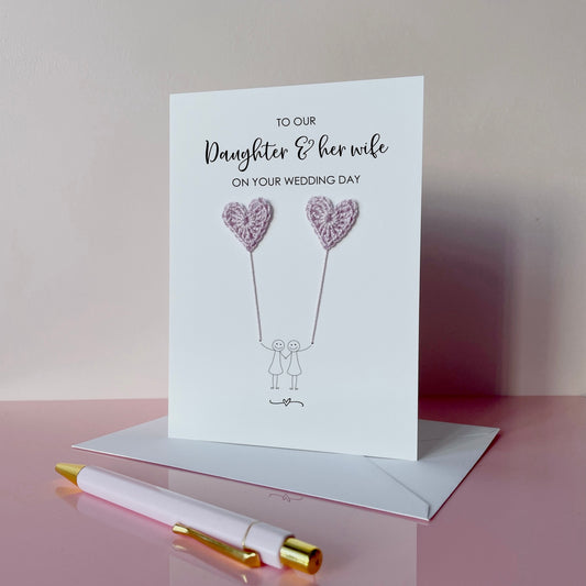 Greeting card with lilac crochet heart balloons and text on a pink surface. The text at the top of the cad reads 'To our Daughter & her wife on your wedding day' 