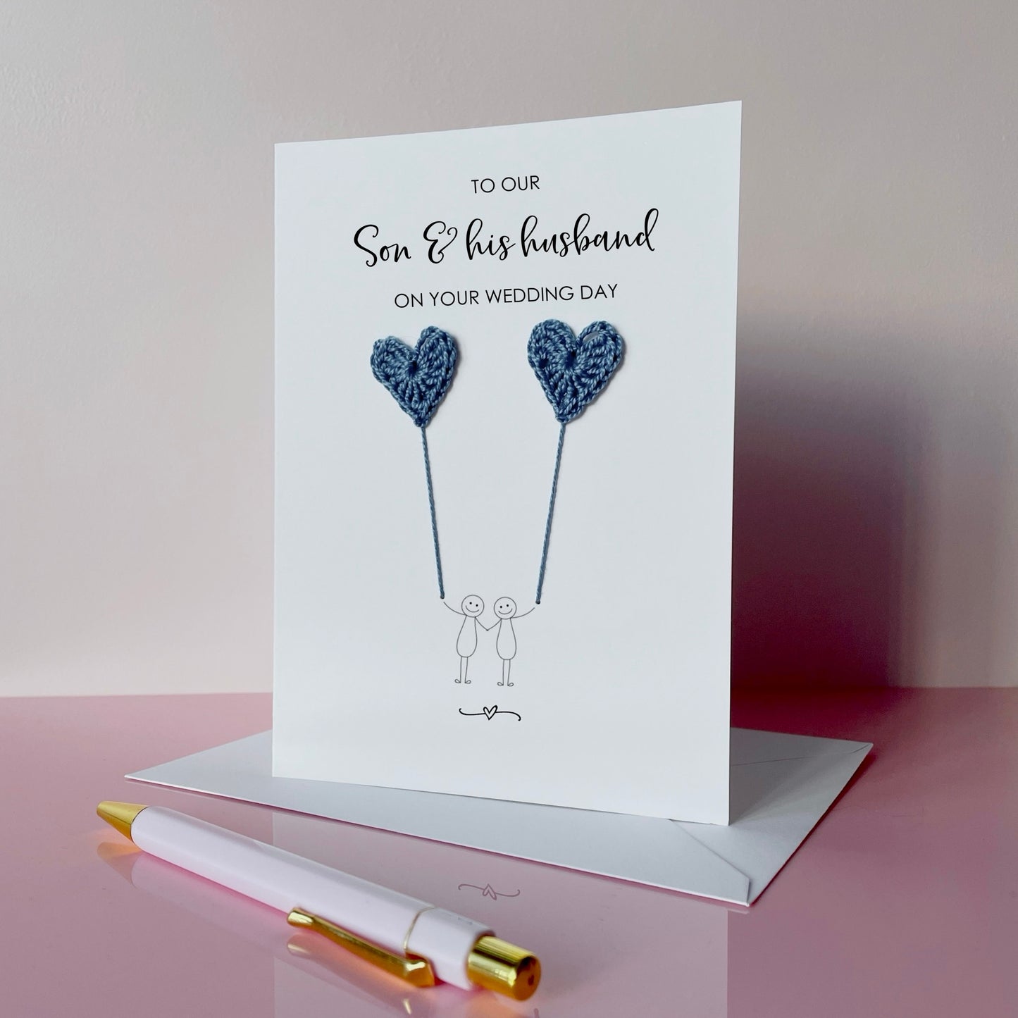A white greeting card with blue crochet heart balloons. The text at the top of the card reads 'To our Son & his husband on your wedding day' 